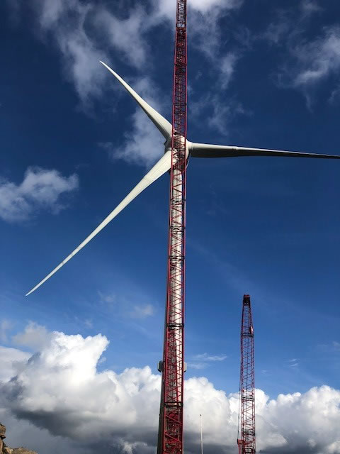 Wind turbine under construction, showcasing a large blade and red cranes against a blue sky. Highlights renewable energy development.