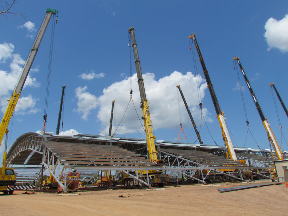 Cranes hoist a large steel structure into place at a construction site under a clear blue sky, highlighting ongoing development work.
